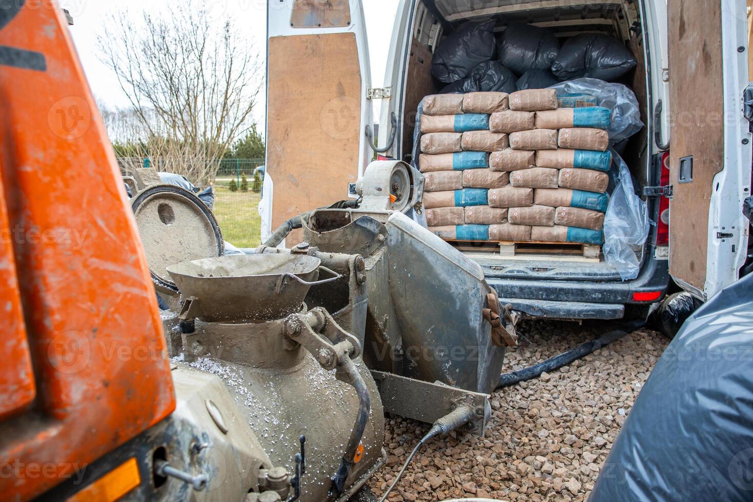 Outdoor construction site featuring a van with open rear doors, cement bags on a pallet, a concrete mixer, and tools on a gravel surface near leafless trees. photo