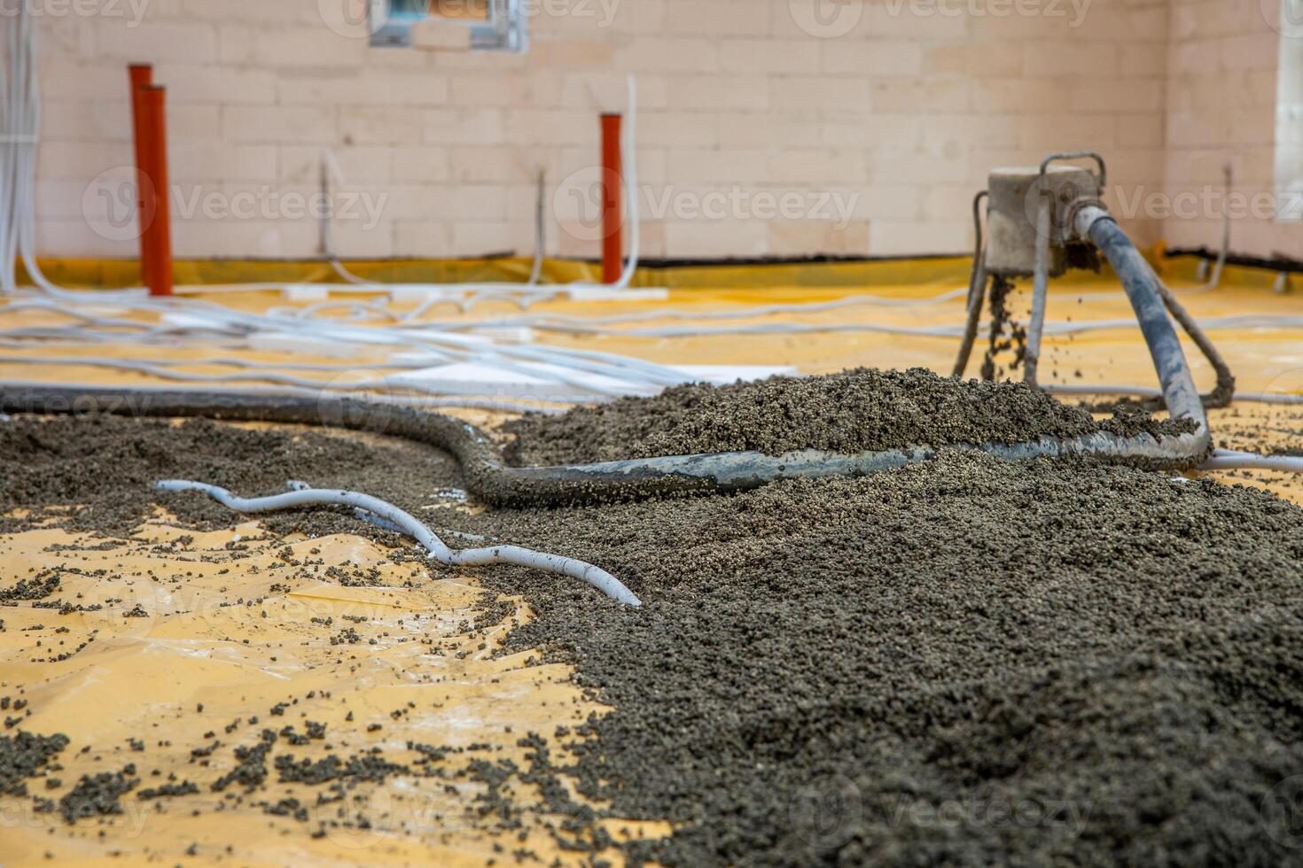 Concrete pouring process on yellow protective sheet in a construction site interior. Red pipes, white tubing, and light brick walls are visible. photo