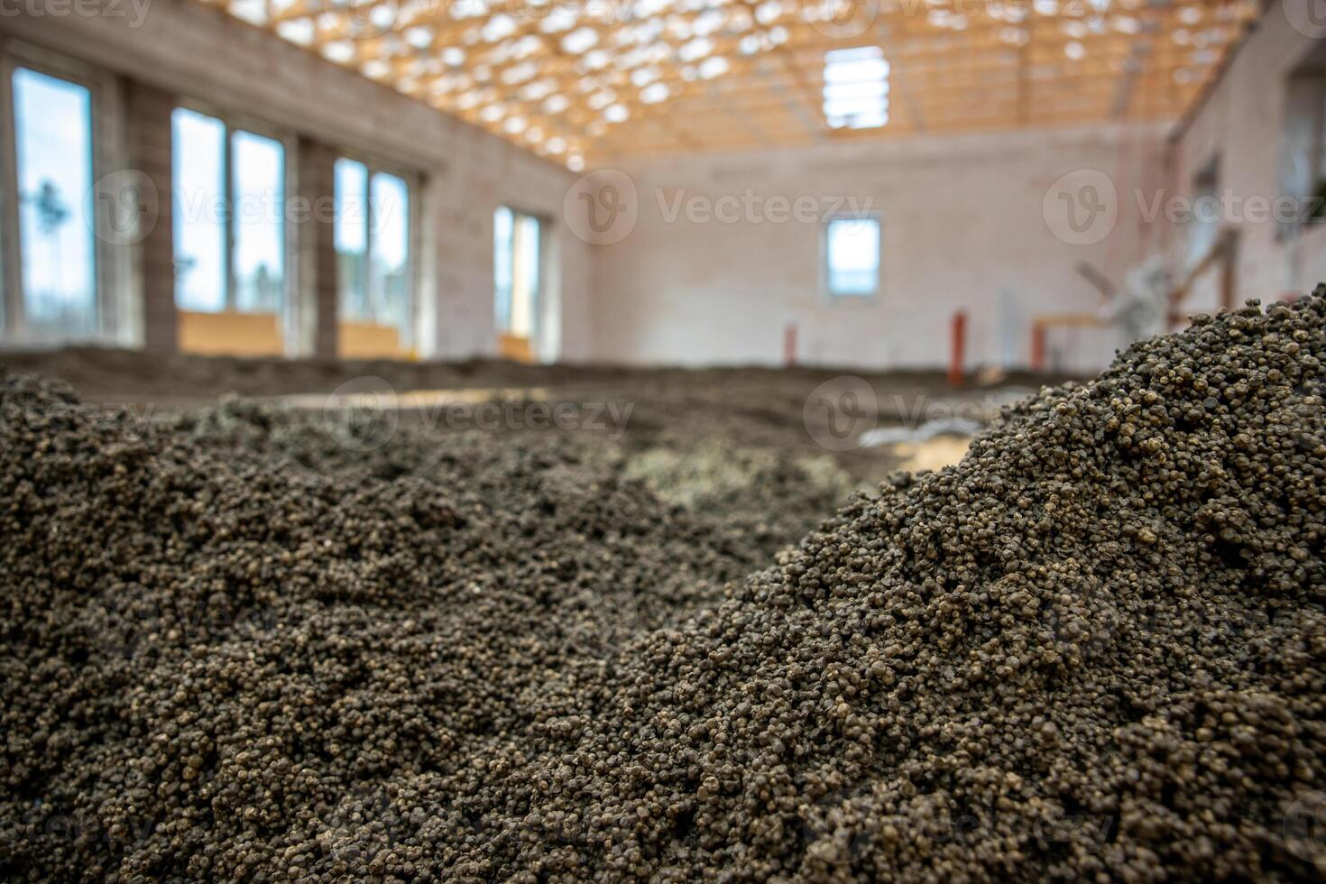 Construction site showing an exposed wooden roof framework, unfinished walls with large window openings, and granular material spread on the floor. photo