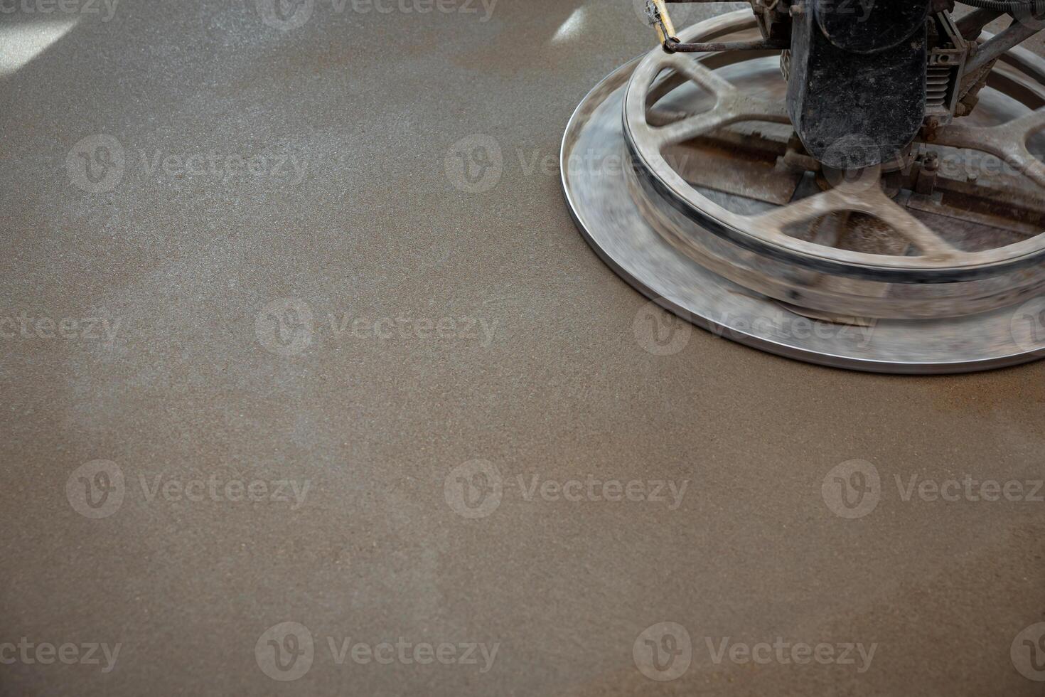 A power trowel machine with a circular metal blade polishes a freshly laid concrete floor, emphasizing texture and sheen under focused lighting. photo