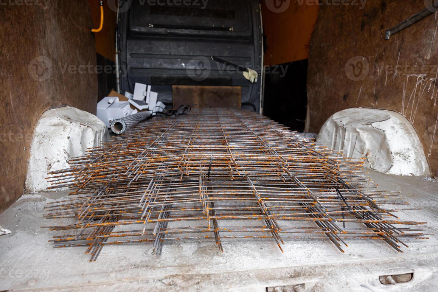 Van interior with stacked metal reinforcement mesh showing rust, wood paneled walls, and scattered construction materials like pipes and papers. photo