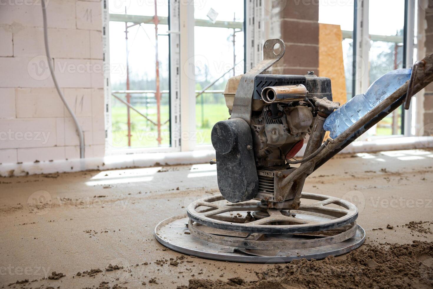 Interior of a construction site showing a power trowel machine on a concrete floor, unfinished walls, large windows, and visible underfloor heating pipes. photo
