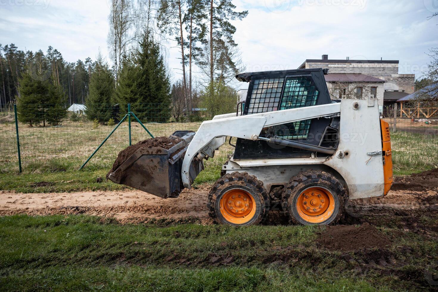A skid steer loader with orange wheels transports soil on a dirt path. The area is fenced, with evergreen trees and a modern flat roof building visible. photo