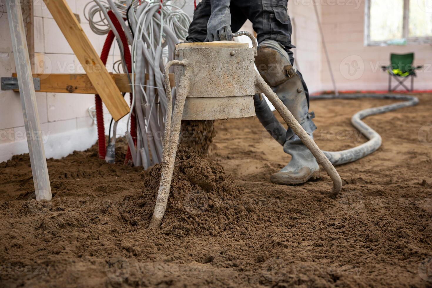 A construction worker pours and levels Estrich concrete over underfloor heating pipes in an unfinished indoor space with visible wiring and materials. photo