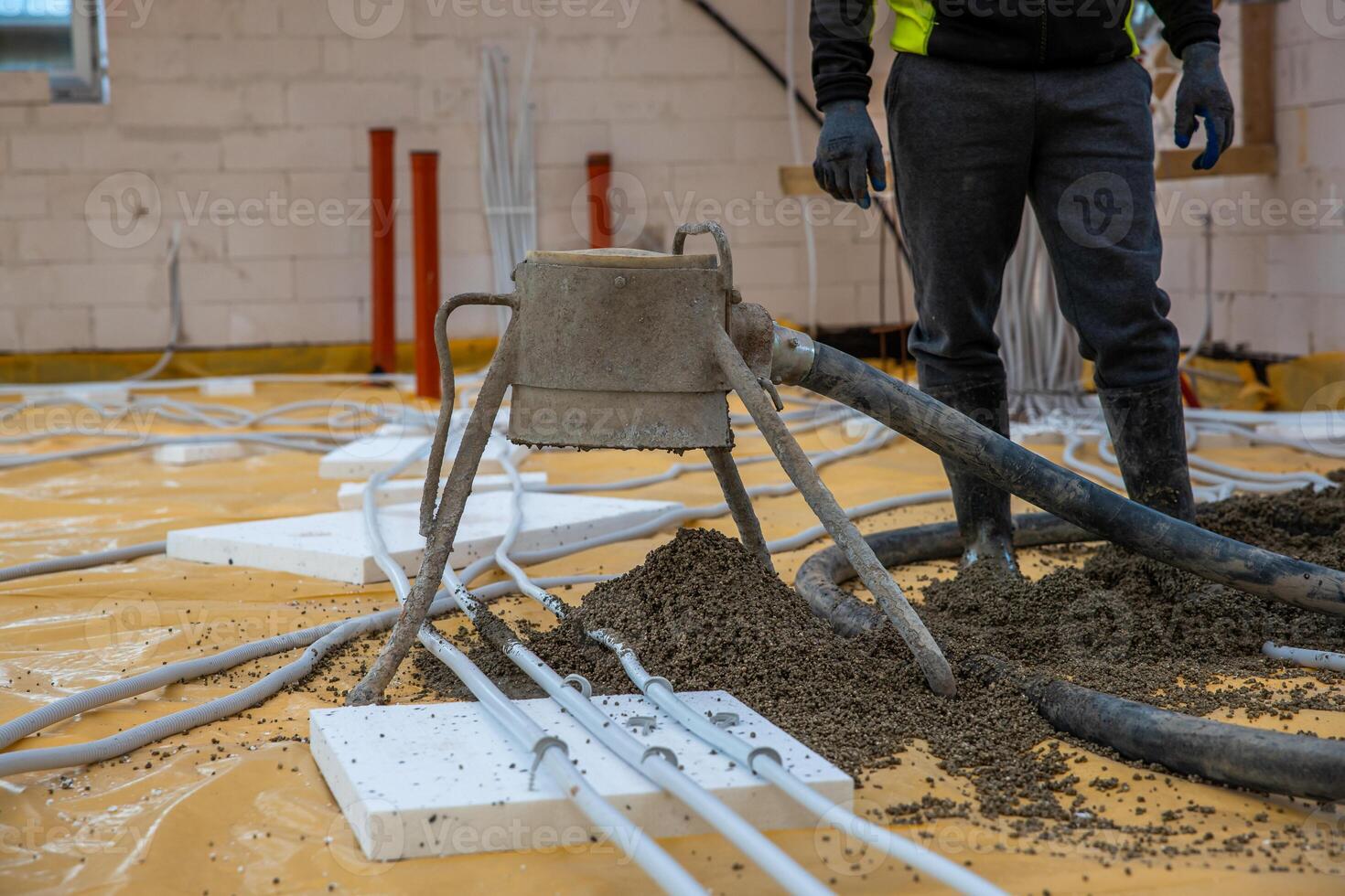 A worker in dark clothing stands near a concrete screed pump on a floor with yellow protective sheets, white insulation panels, and visible piping. photo