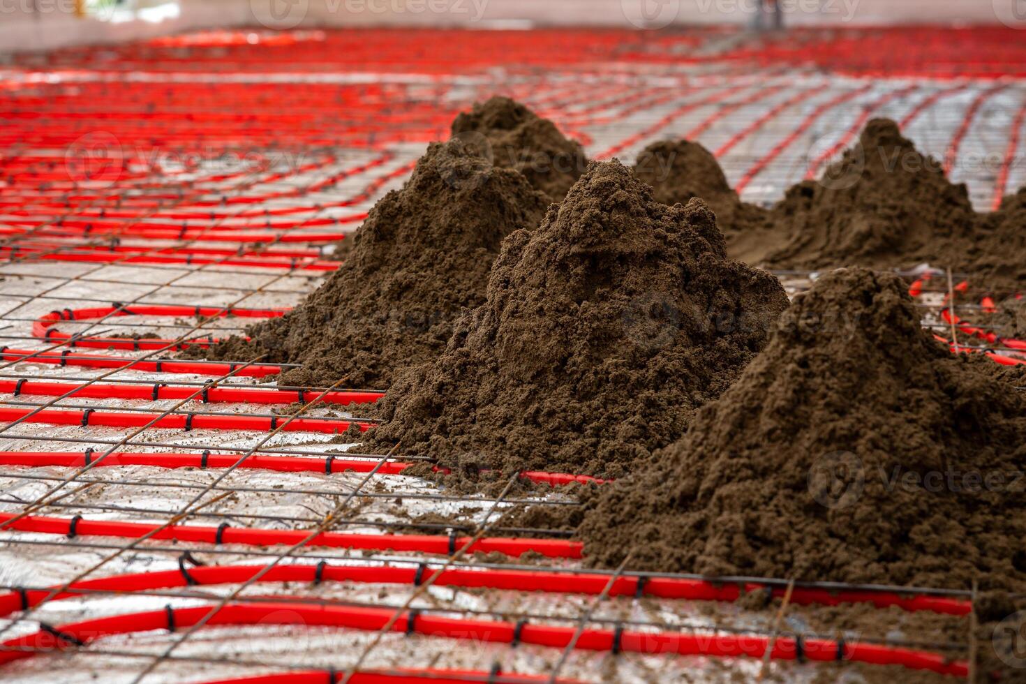 Red underfloor heating pipes in a grid pattern secured with metal mesh, with piles of Estrich concrete ready for spreading and leveling. photo