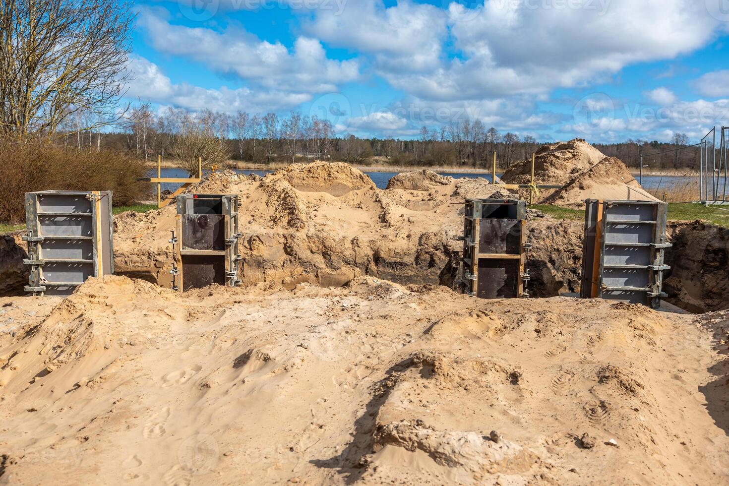 Construction site featuring metal framework in sandy soil, piles of sand, and a body of water bordered by sparse trees under a partly cloudy sky. photo