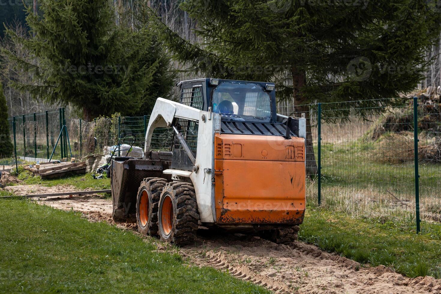 An orange and white skid steer loader rests on a dirt path with its bucket down. The area features tire tracks, disturbed soil, and tall evergreen trees. photo