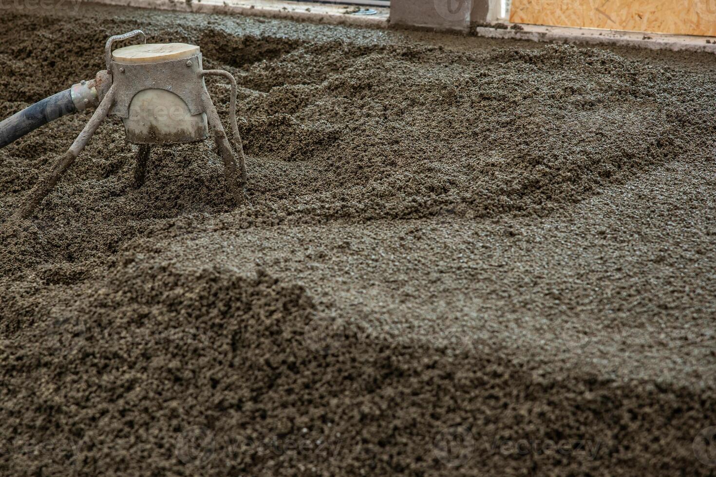 Freshly poured concrete floor in an indoor construction site, with a leveling tool connected to a hose in use. Partially visible wall and window frame. photo