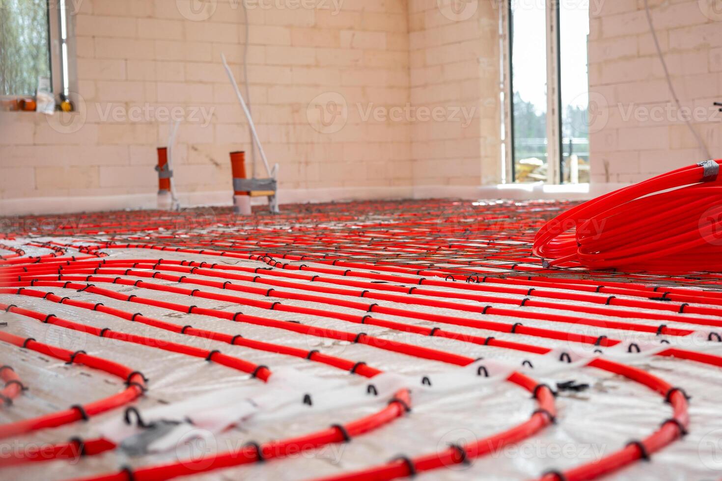 Interior of a construction site showing red underfloor heating pipes in a grid pattern over reflective insulation, with light brick walls and window openings. photo