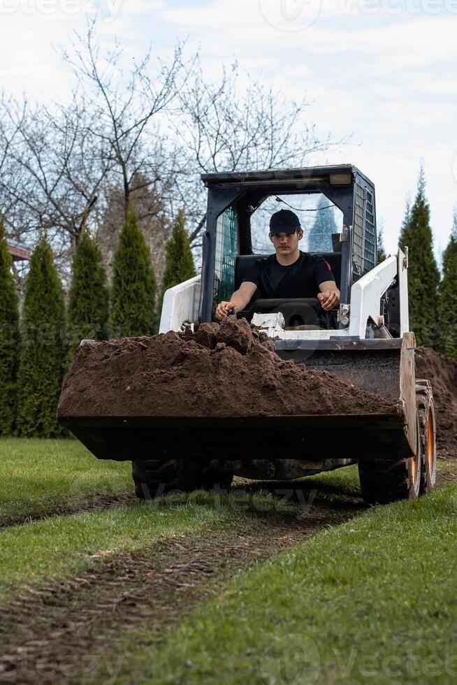 A man in a black cap and shirt operates a skid steer loader carrying soil across grass. Tracks are visible, with conifer and deciduous trees in the background. photo