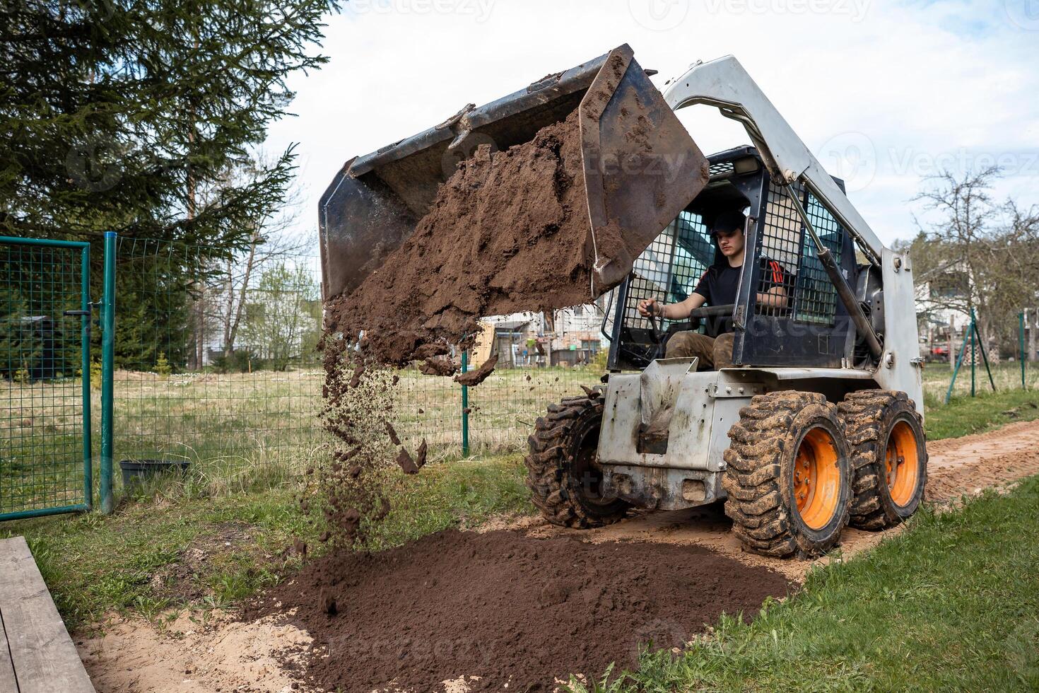 A skid steer loader with orange wheels dumps soil onto a pathway. The operator works in a fenced area with grass, sparse trees, and distant buildings. photo
