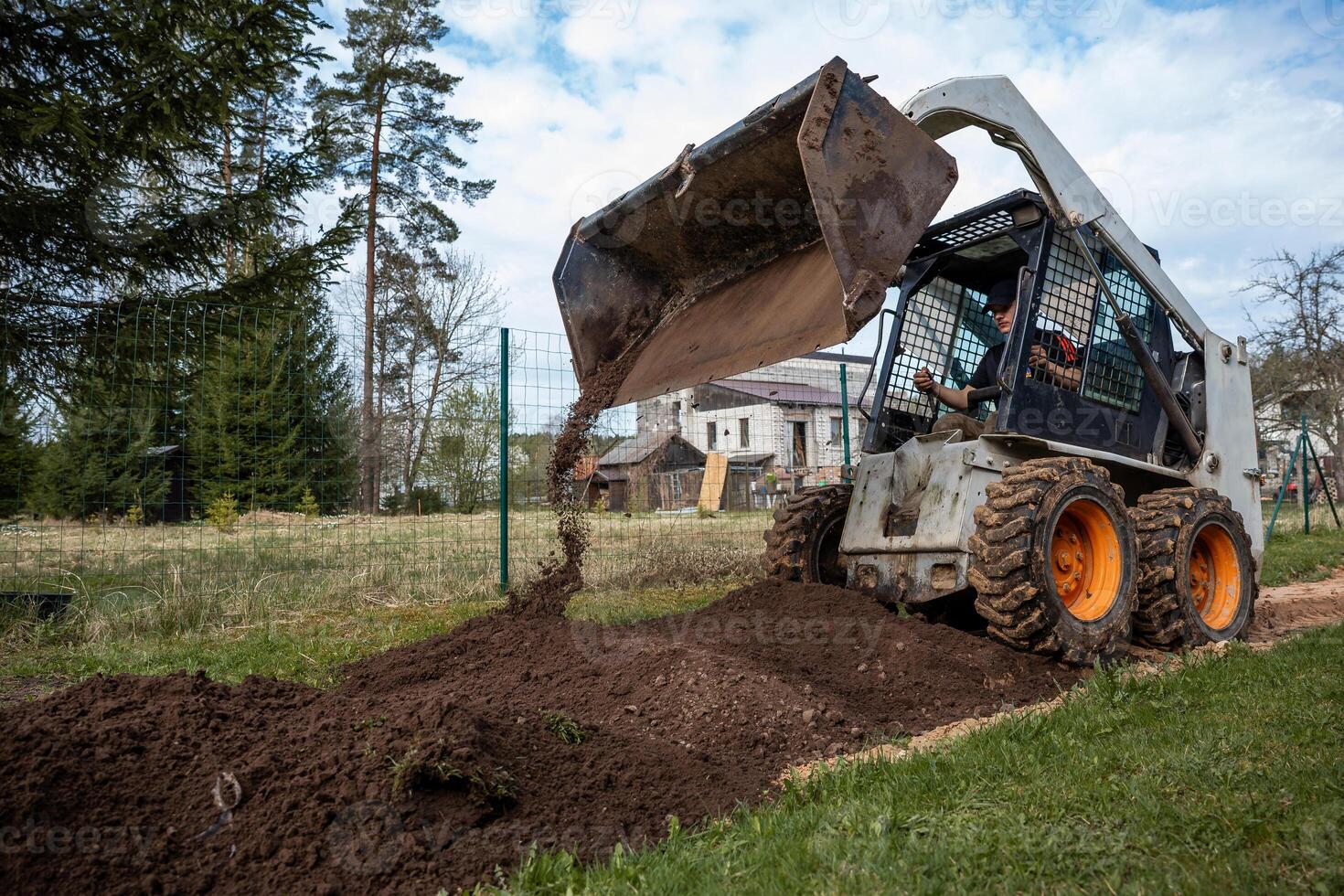 A skid steer loader with orange wheels pours soil near a white house with scaffolding. Wire fence, tall trees, and grassy areas surround the site. photo