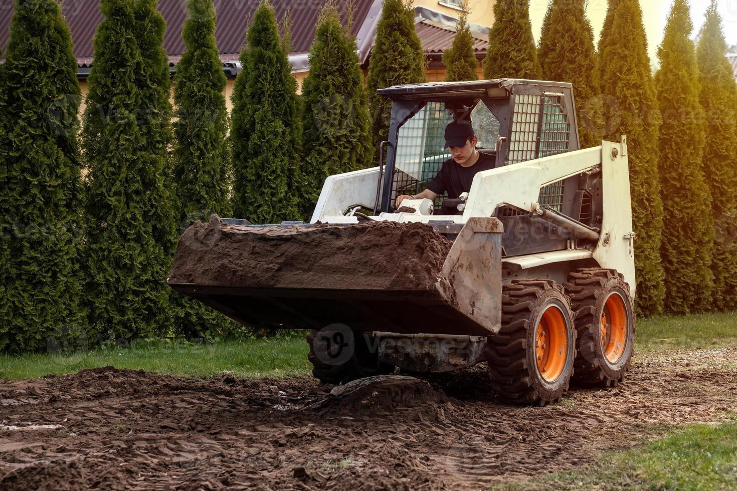 A skid steer loader with a raised bucket moves soil near tall evergreen trees. A building with a sloped roof is partially visible in the setting. photo