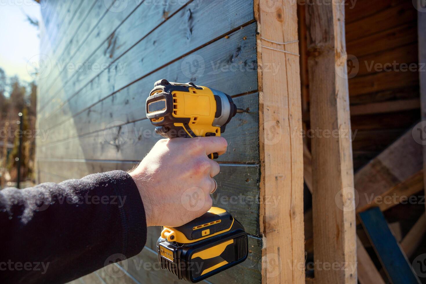 A person uses a yellow and black cordless drill to secure dark wooden planks. The scene includes wooden elements and natural light in an outdoor setting. photo
