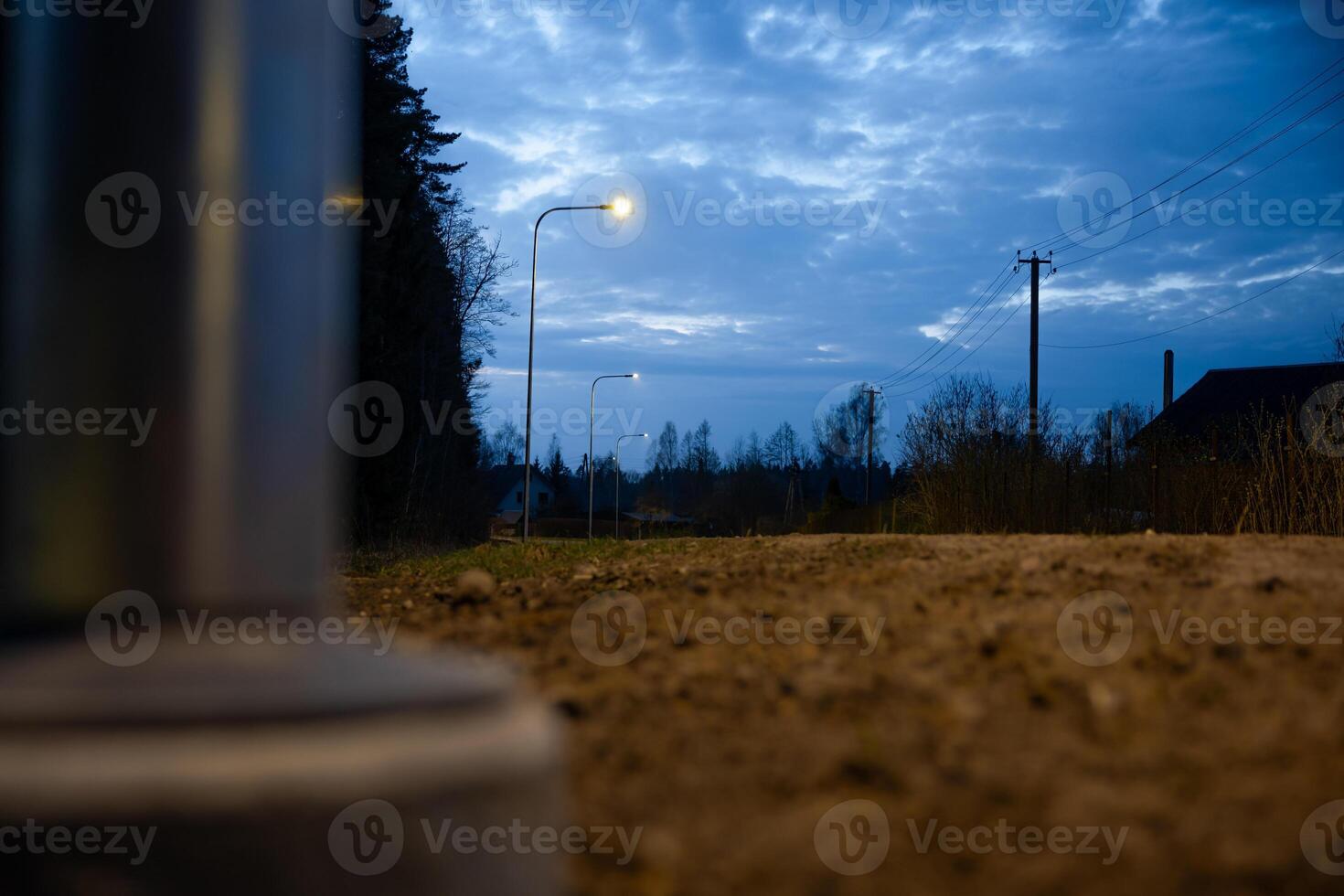 A dirt pathway lit by streetlights under a deep blue evening sky, with power lines, a utility pole, tree silhouettes, and a blurred metallic object in view. photo