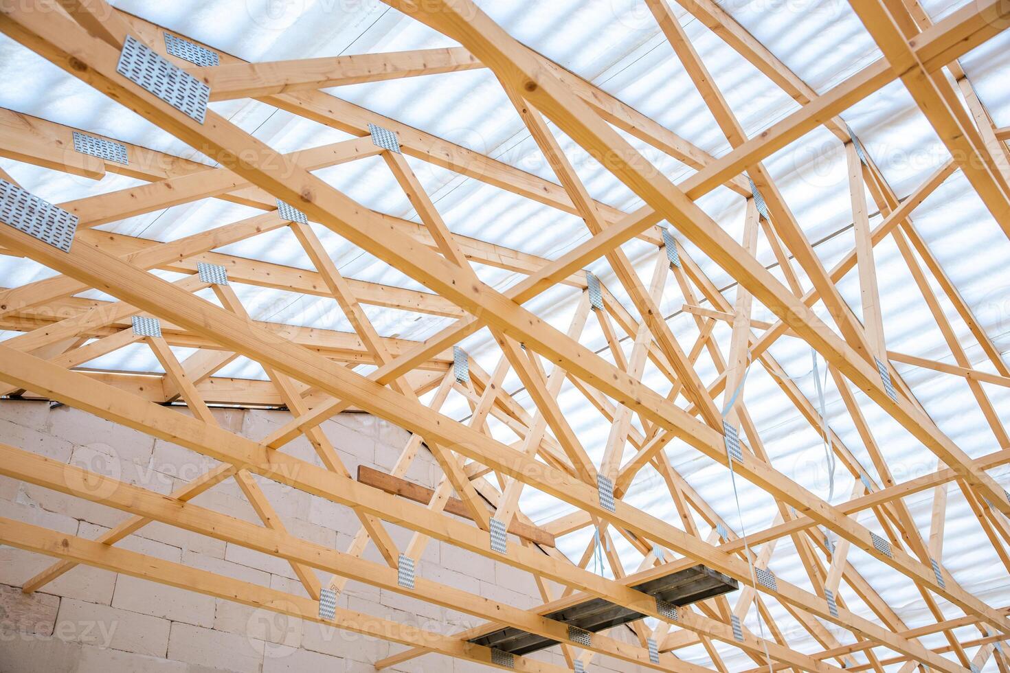 Interior view of a construction site featuring wooden trusses in a geometric pattern, a translucent roof, and light colored concrete block walls. photo