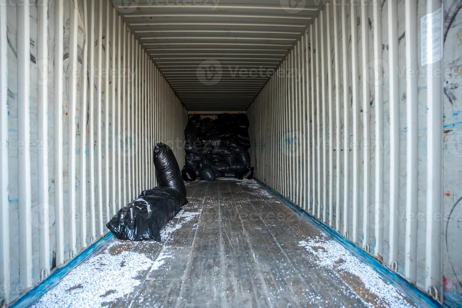 White walled shipping container with ribbed structure, wooden floor, black plastic bags at the far end, and scattered white particles on the floor. photo