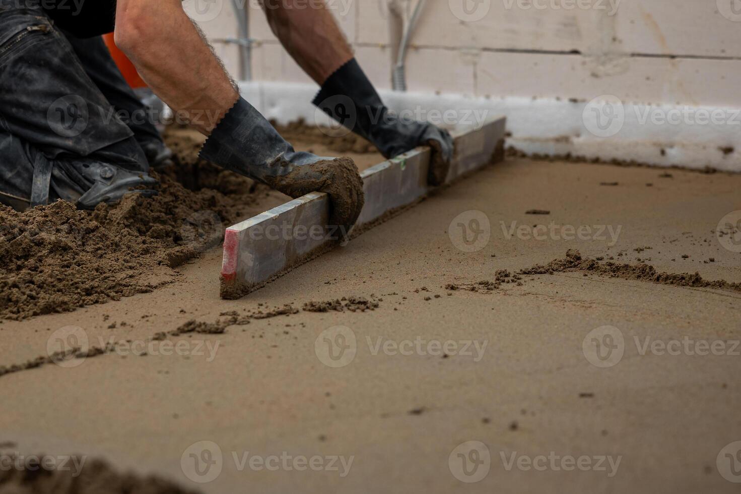 A worker kneels on wet concrete, leveling it with a straightedge tool. Underfloor heating pipes are visible, with unfinished walls and wiring around. photo
