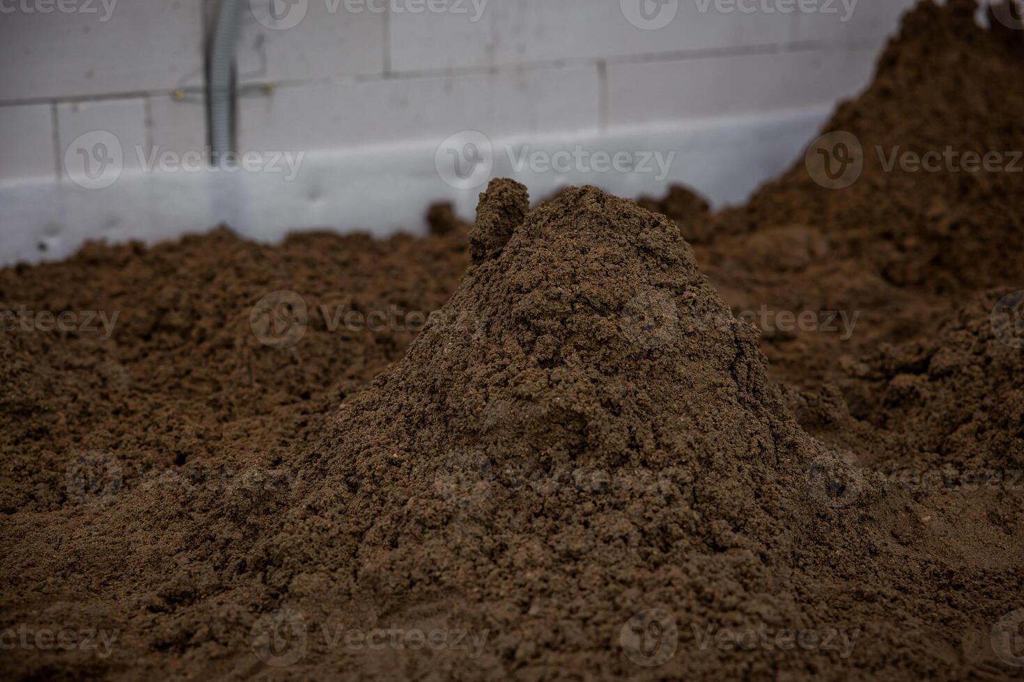 Indoor construction site with a pile of brown soil, a white wall with seams, and exposed piping, prepared for underfloor heating or concrete work. photo