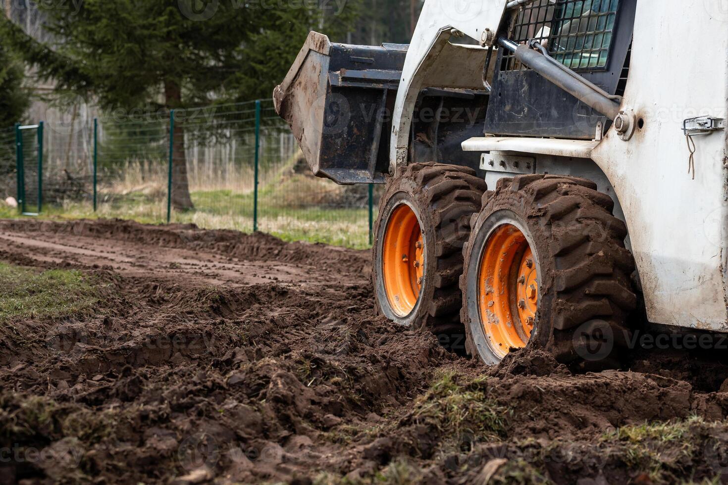 A skid steer loader with orange wheels sits on a muddy dirt path, its bucket raised. A green metal fence and sparse vegetation are visible nearby. photo