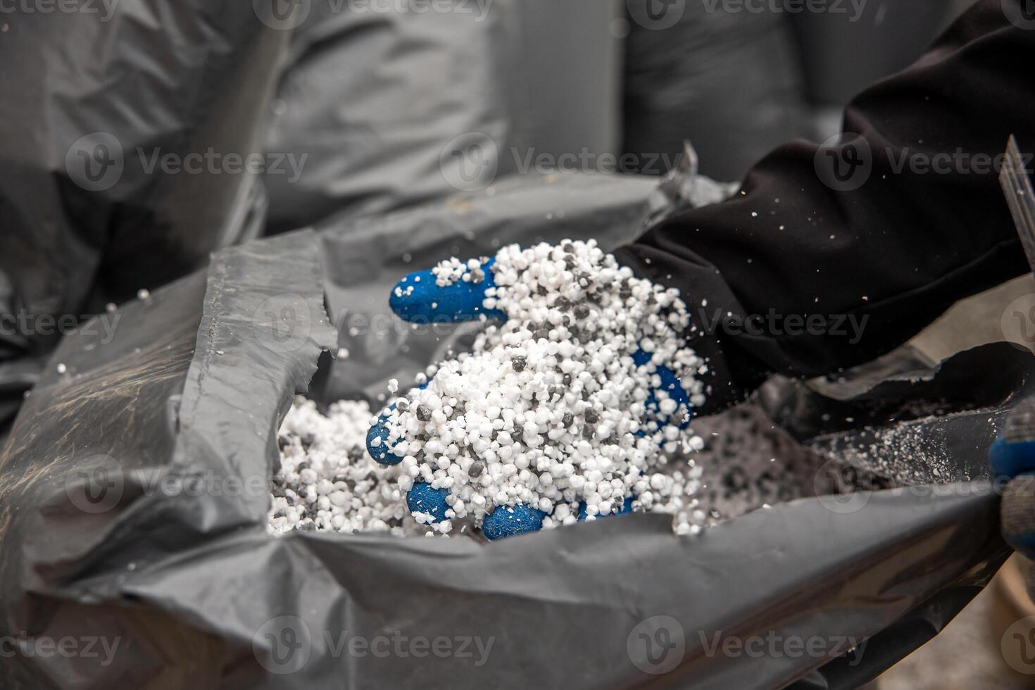 A close up of a gloved hand holding small white granules, likely fertilizer, scooped from a large gray bag in an industrial or agricultural setting. photo