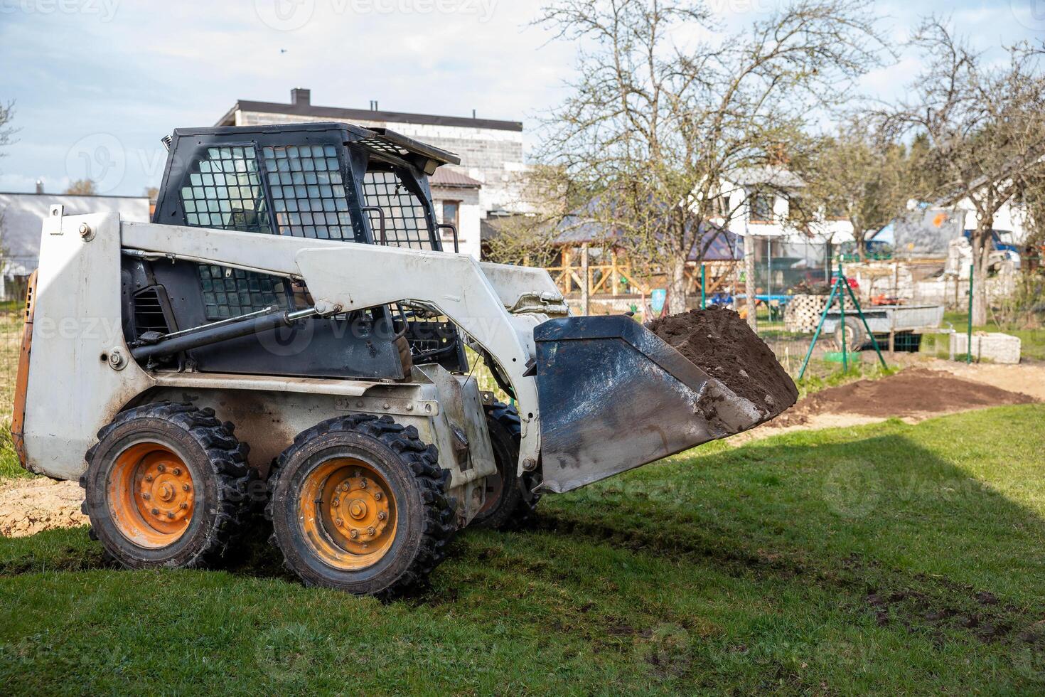A skid steer loader with orange wheels carries soil in its raised bucket on a grassy surface. Nearby are houses, a swing set, and leafless trees. photo