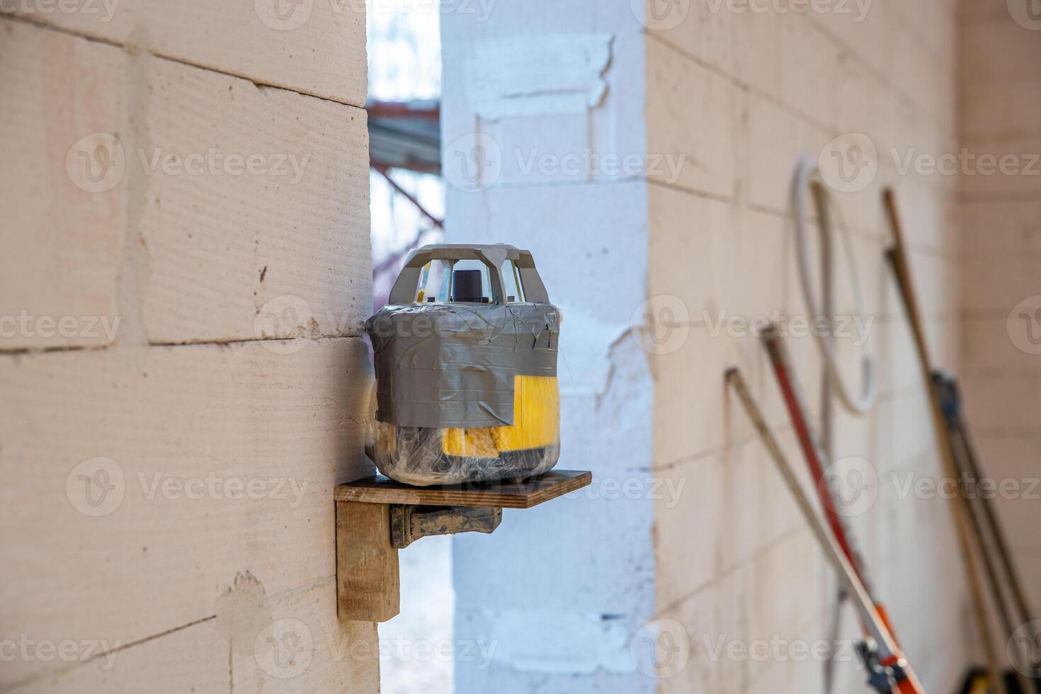 A makeshift device wrapped in duct tape sits on a wooden shelf against a light brick wall, surrounded by tools and an unfinished workshop environment. photo