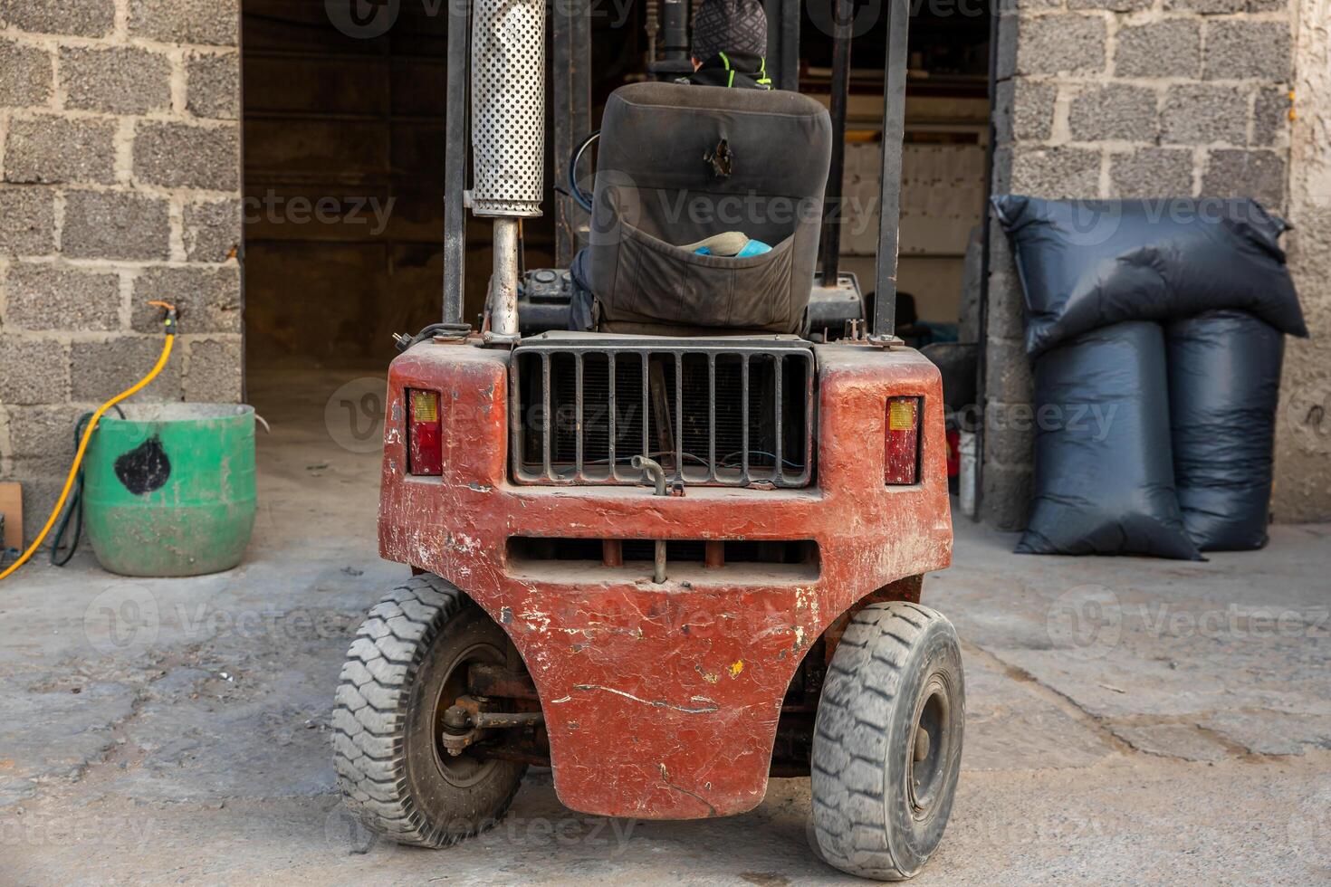 A red forklift with visible wear stands near a stone walled structure with an open doorway. A green container and black bags are placed nearby on concrete ground. photo