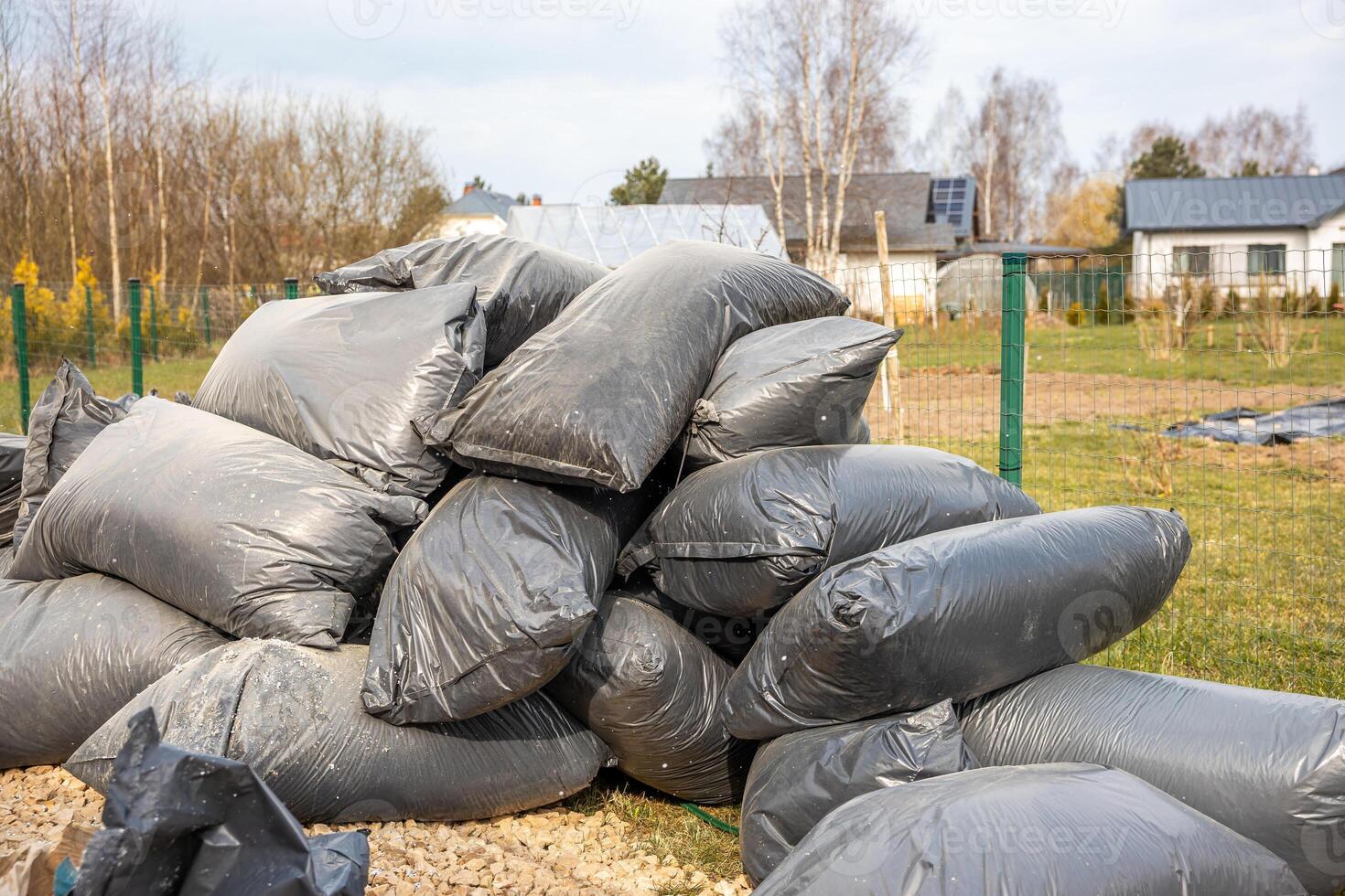A pile of black plastic bags rests on a gravel surface, enclosed by a green wire fence. Nearby are residential houses, a greenhouse, and leafless trees. photo