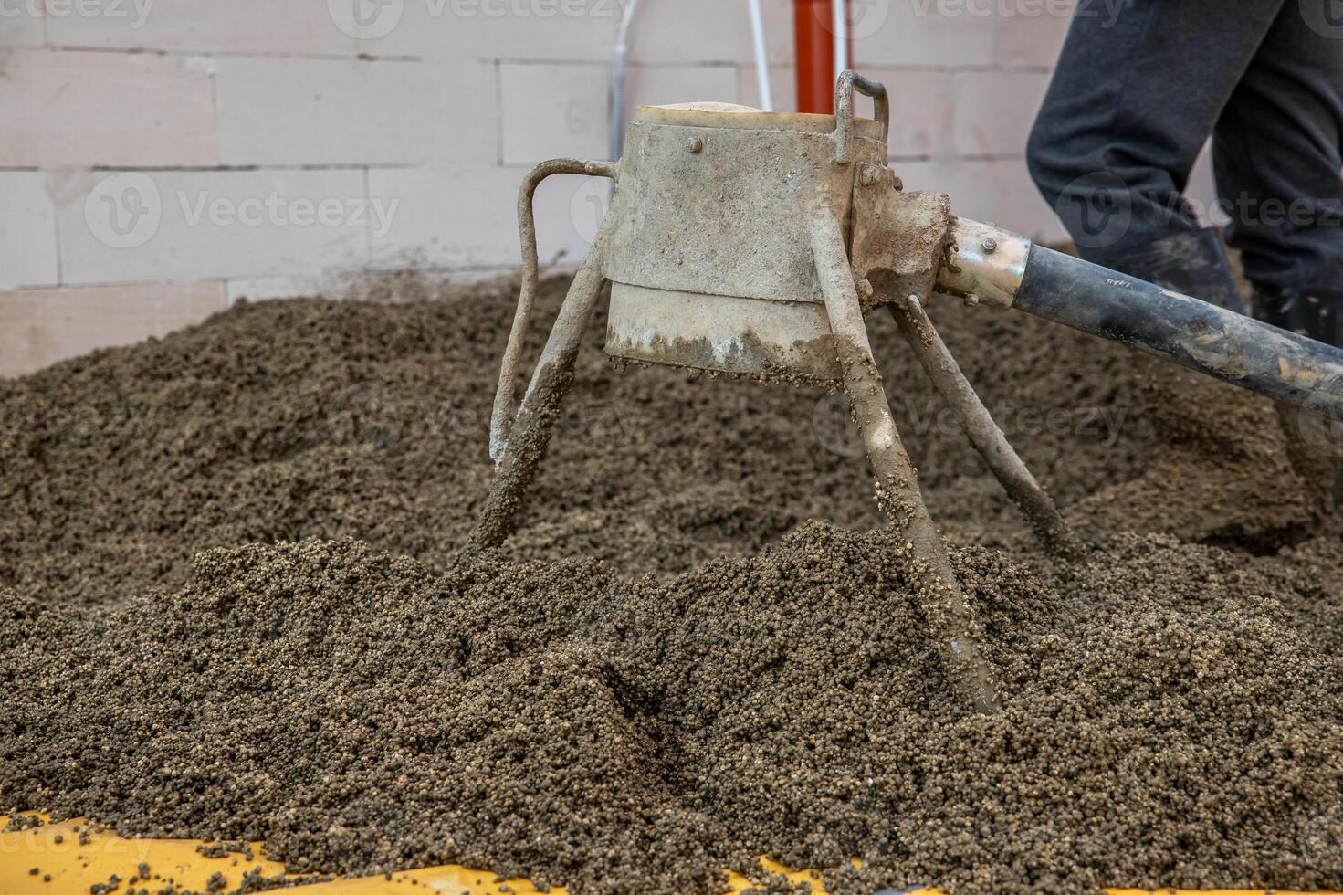 A concrete screed tool with a tripod structure and hose rests on lightweight aggregate concrete. A worker stands nearby near a white brick wall and red pipe. photo