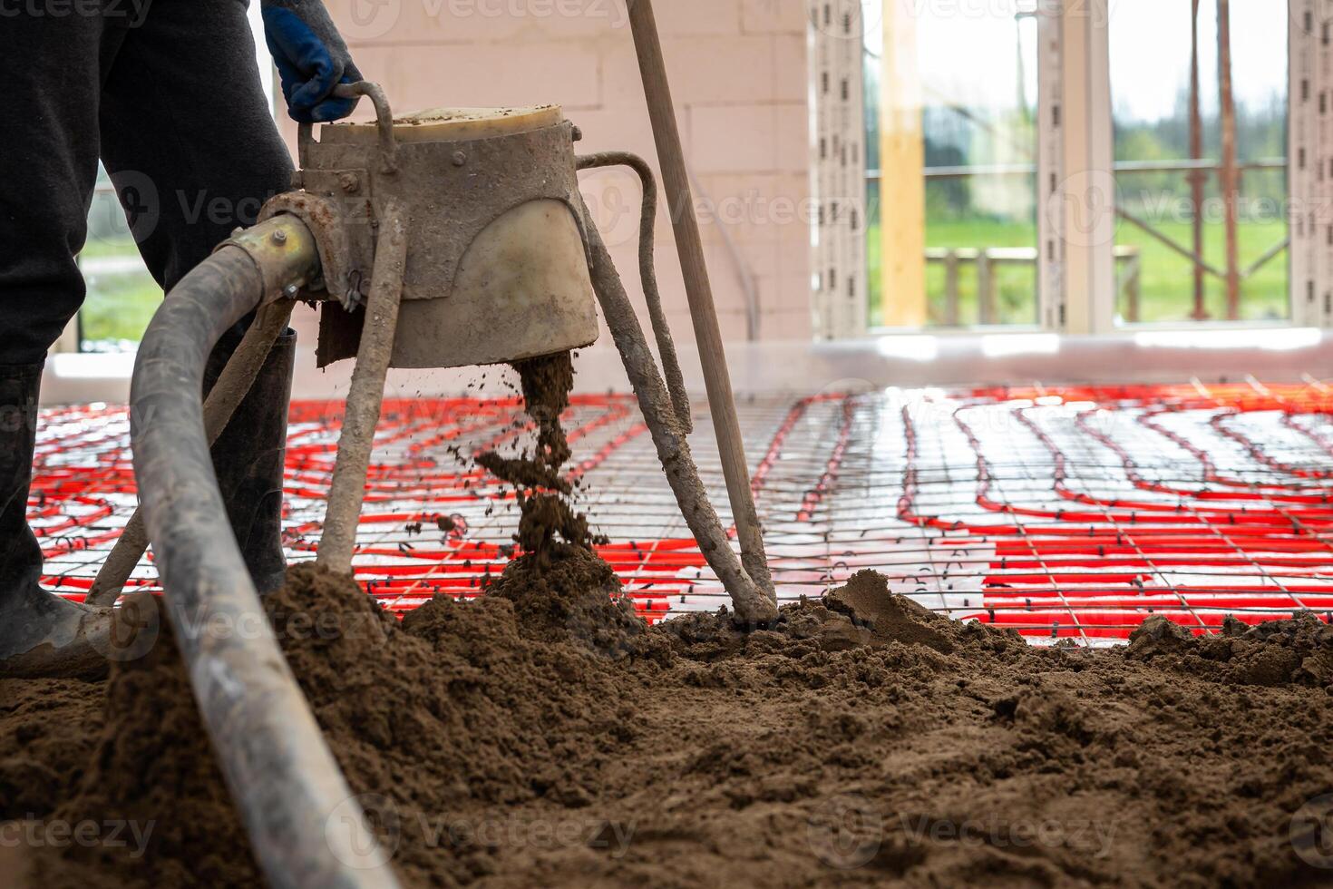 Red heating pipes are arranged on the floor as a worker pours screed concrete using a pump inside a building under construction with open windows. photo