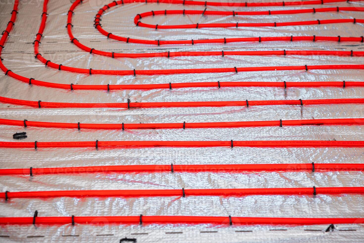 Bright red underfloor heating pipes arranged in loops, secured with black fasteners on a silver insulation layer during the installation phase. photo