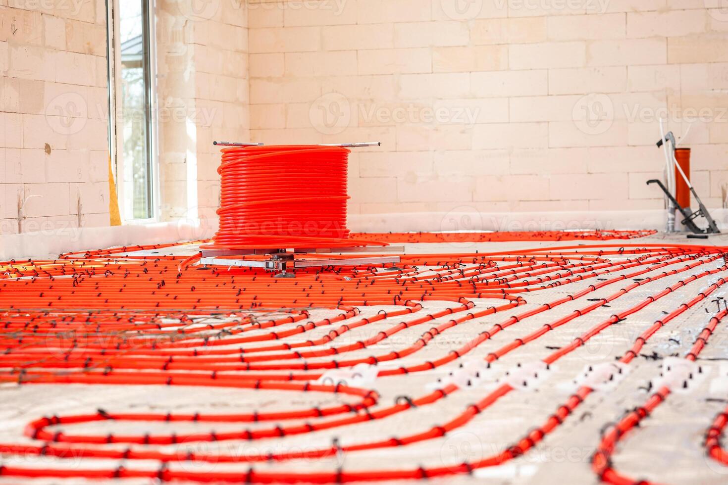 Construction site interior with red underfloor heating pipes laid in a pattern, light brick walls, a window, and a pipe installation tool in the corner. photo