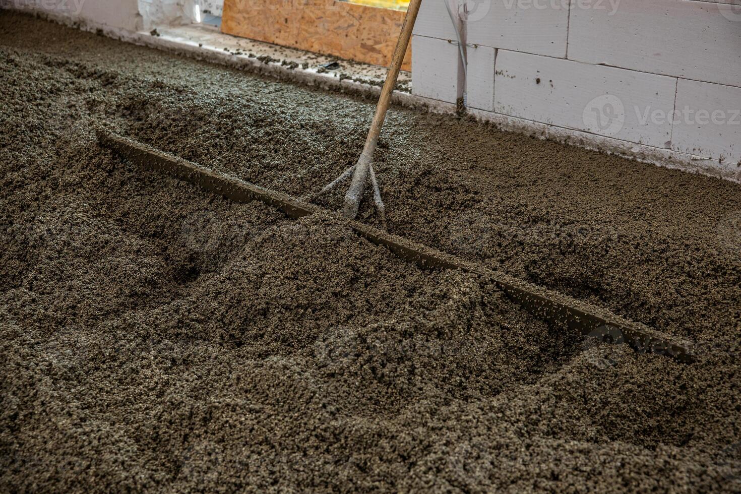 An indoor construction site shows wet concrete being leveled with a rake like tool. Unfinished white block walls and a wooden panel are visible. photo