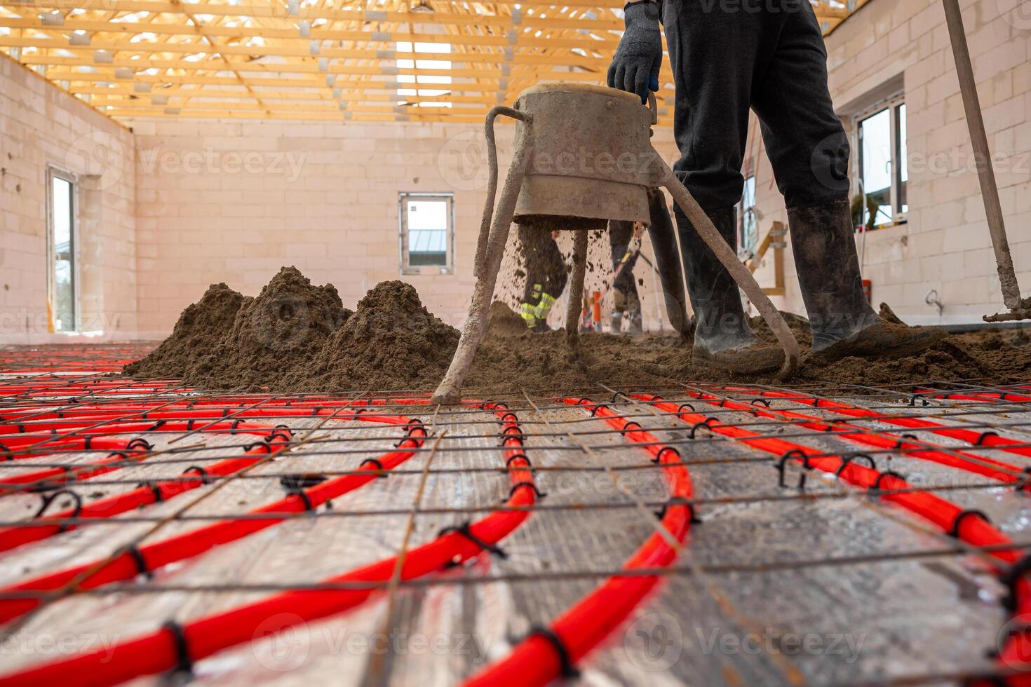 A construction worker installs red underfloor heating pipes in a systematic layout and pours Estrich concrete in a building with unfinished walls and roof frame. photo