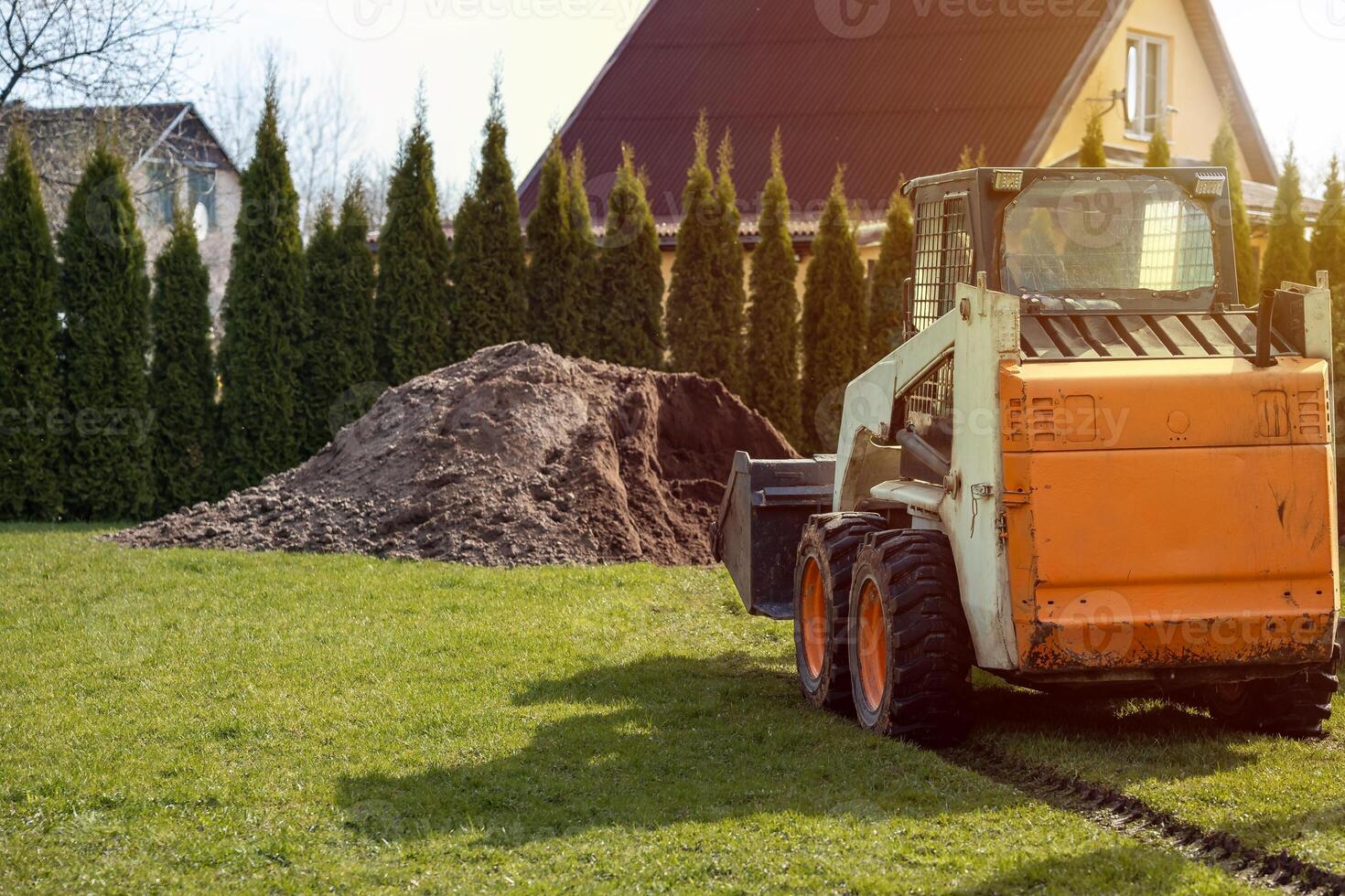 An orange and white skid steer loader sits on grass with tire tracks nearby. A soil pile and tall evergreen trees partially obscure a yellow house. photo