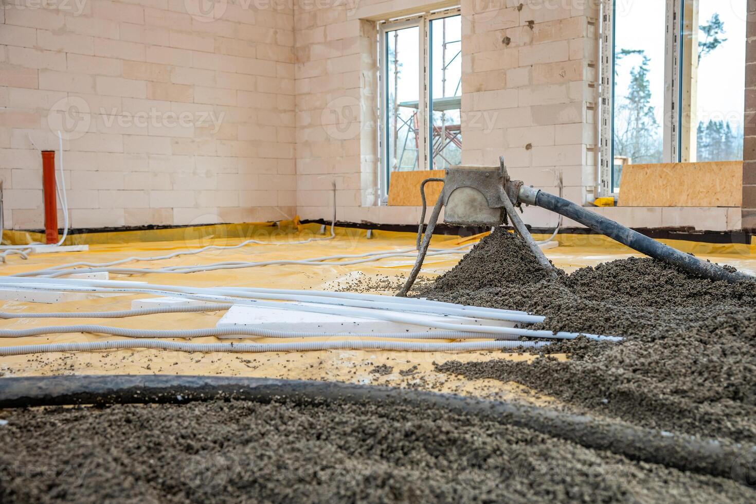 An under construction interior with light brick walls, concrete pouring via a hose, yellow floor covering, white pipes, and large windows with scaffolding outside. photo