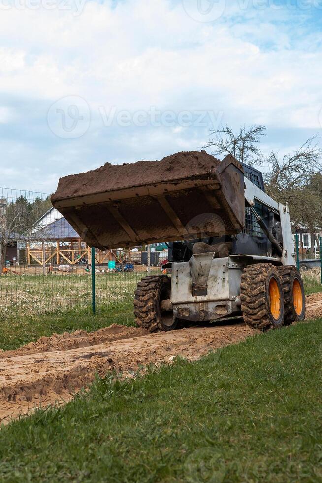 A skid steer loader with a raised bucket of soil sits on a muddy dirt path, bordered by a wire fence and a wooden structure under an overcast sky. photo