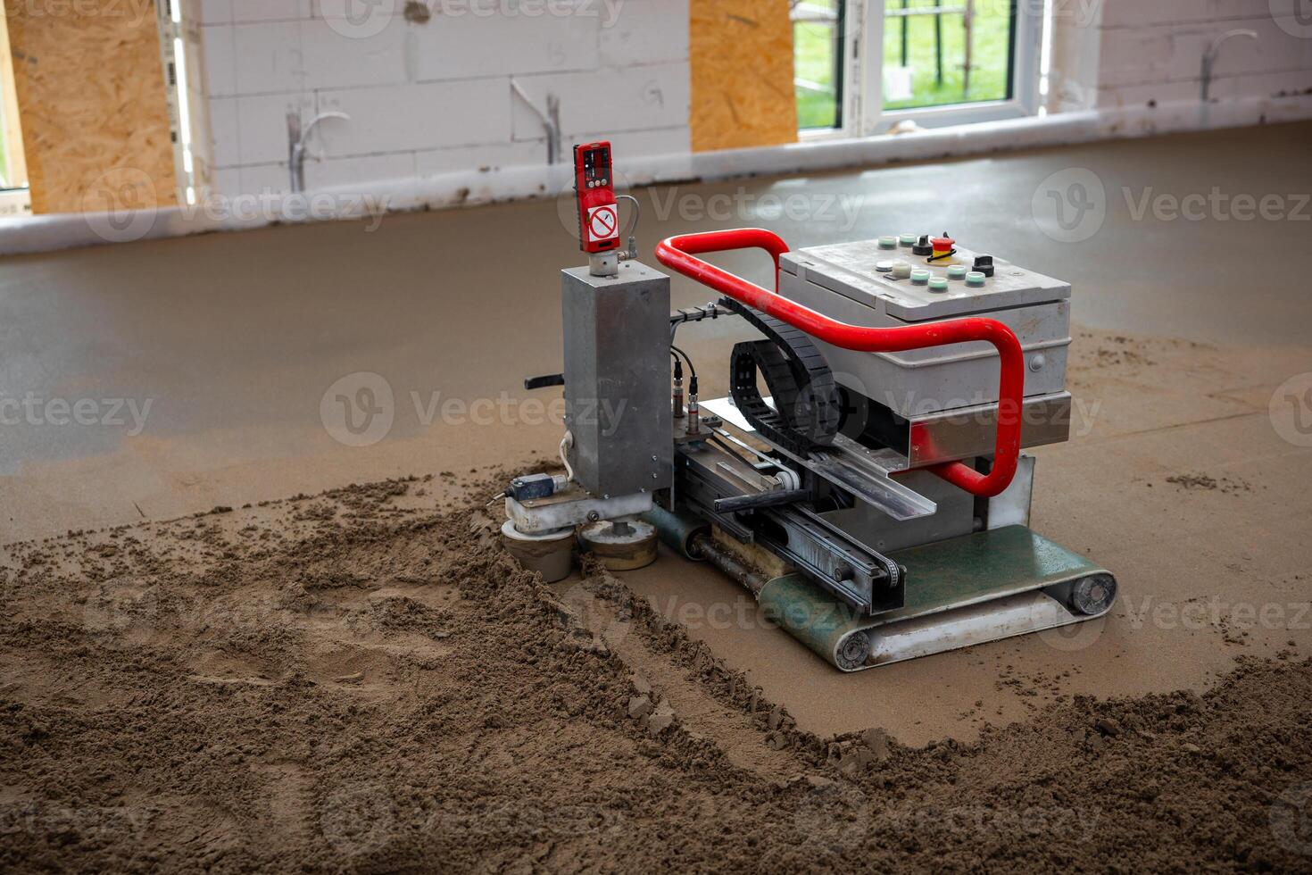 A machine with a red handle levels screed concrete on a construction site floor. Exposed white brick walls and large windows provide natural light. photo