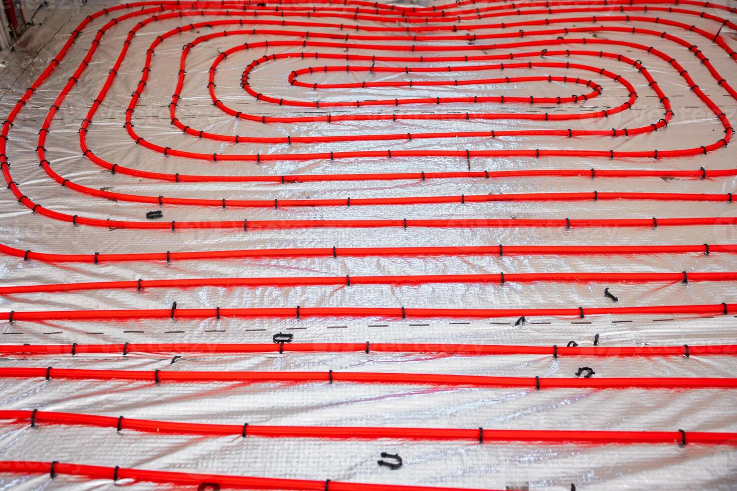 Construction site floor with red underfloor heating pipes arranged in loops over a reflective insulation layer, secured with black fasteners. photo