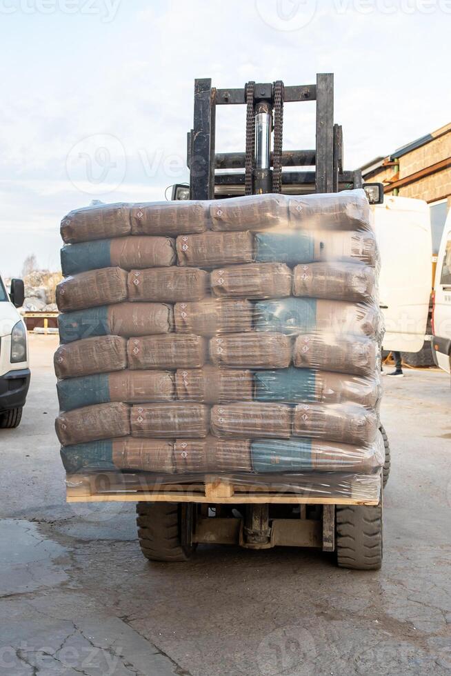 A forklift carries a pallet of clear wrapped bags on paved ground near a white van and brick building, indicating active material handling. photo
