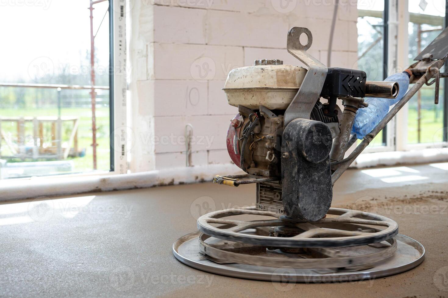 A power trowel machine smooths fresh concrete flooring inside a white brick structure with large windows and visible scaffolding outside. photo
