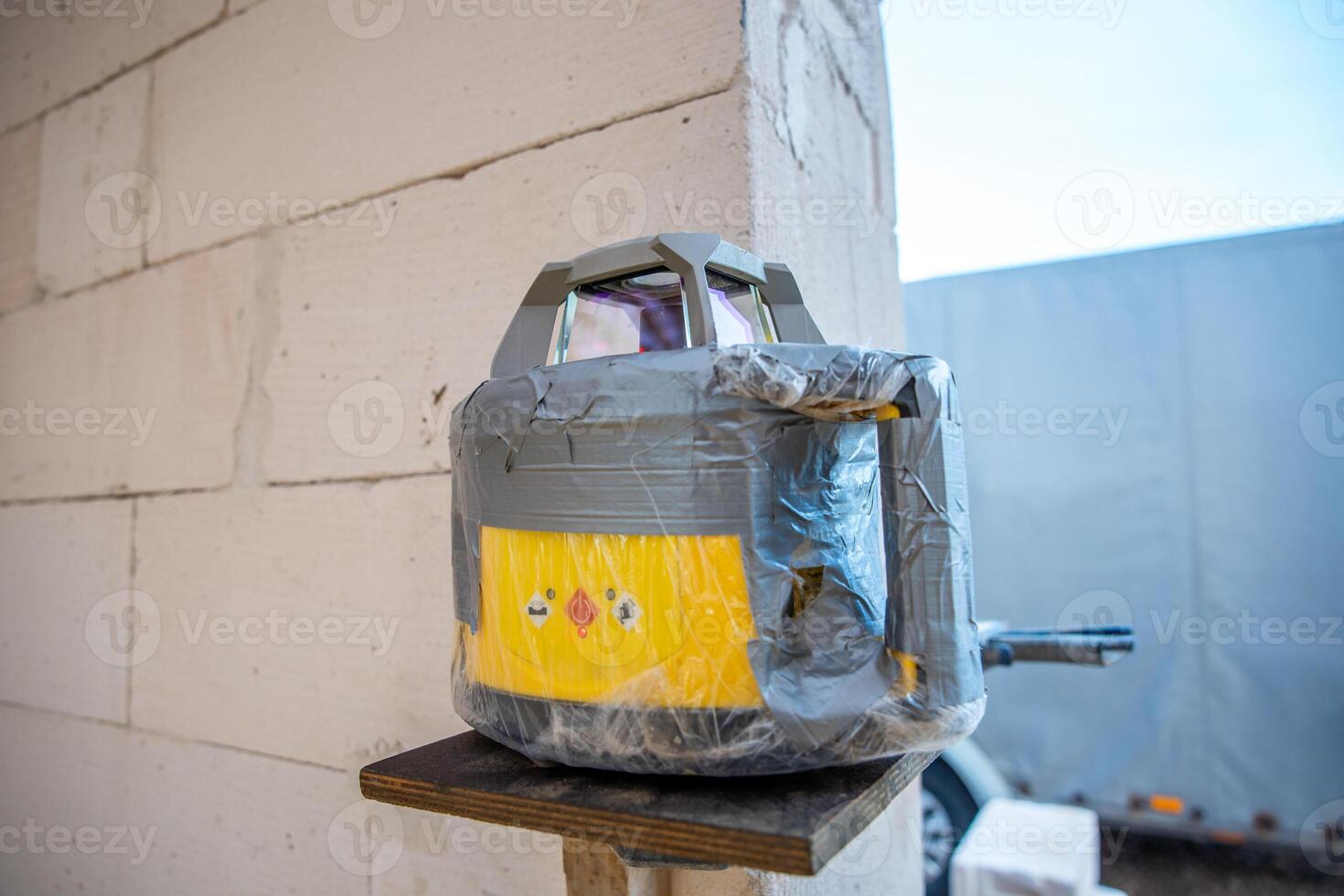 A yellow and gray surveying device wrapped in plastic and duct tape is mounted on a wooden platform near a light brick wall. A white vehicle and gray structure are visible. photo