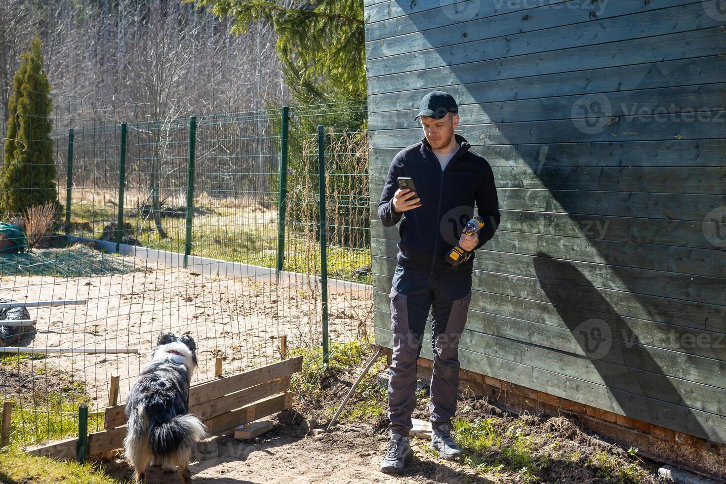 A man in dark clothing stands by a green wooden structure holding a smartphone and bottle. A black and white dog walks near a fenced dirt path with sparse trees. photo