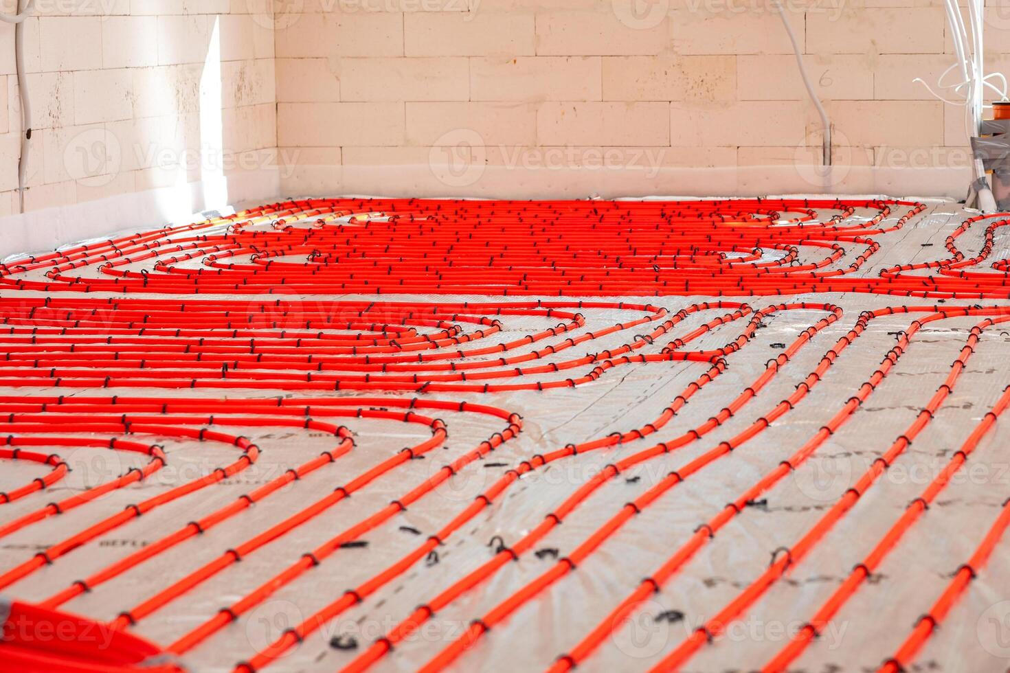 Interior of a construction site showing red underfloor heating pipes arranged in loops on a base layer, with light brick walls and visible electrical conduits. photo