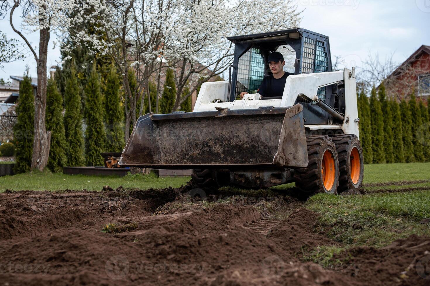 A skid steer loader with orange wheels works on tilled soil. A person in a black cap operates it near flowering trees, hedges, and a red roofed house. photo