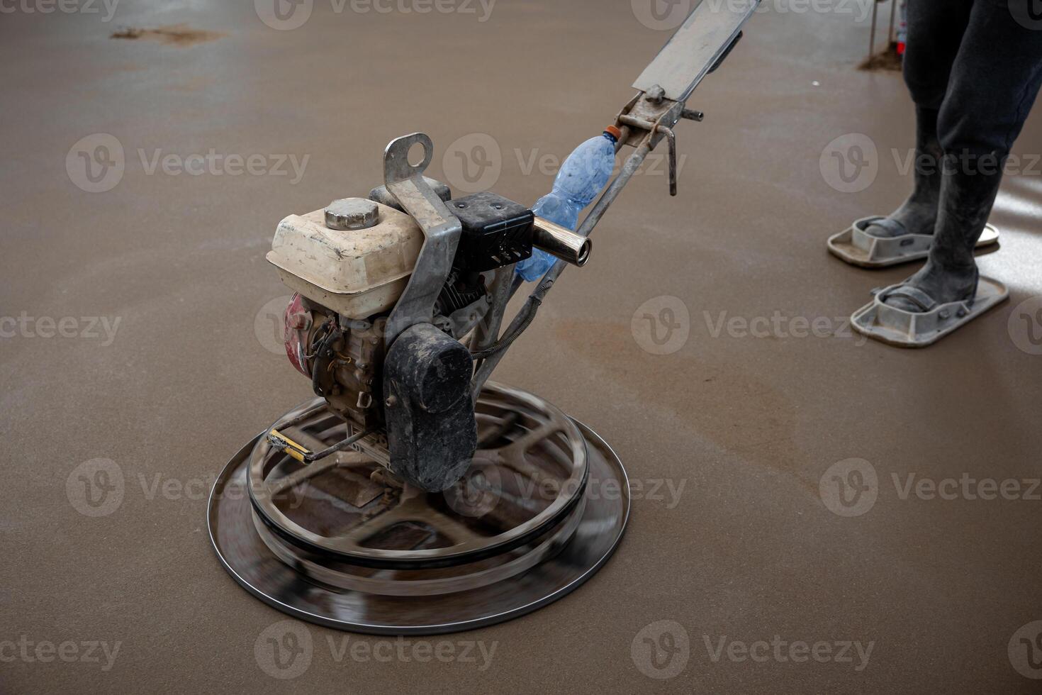 A construction worker operates a power trowel machine indoors, smoothing a freshly laid concrete floor. Protective footwear and active trowel blades are visible. photo