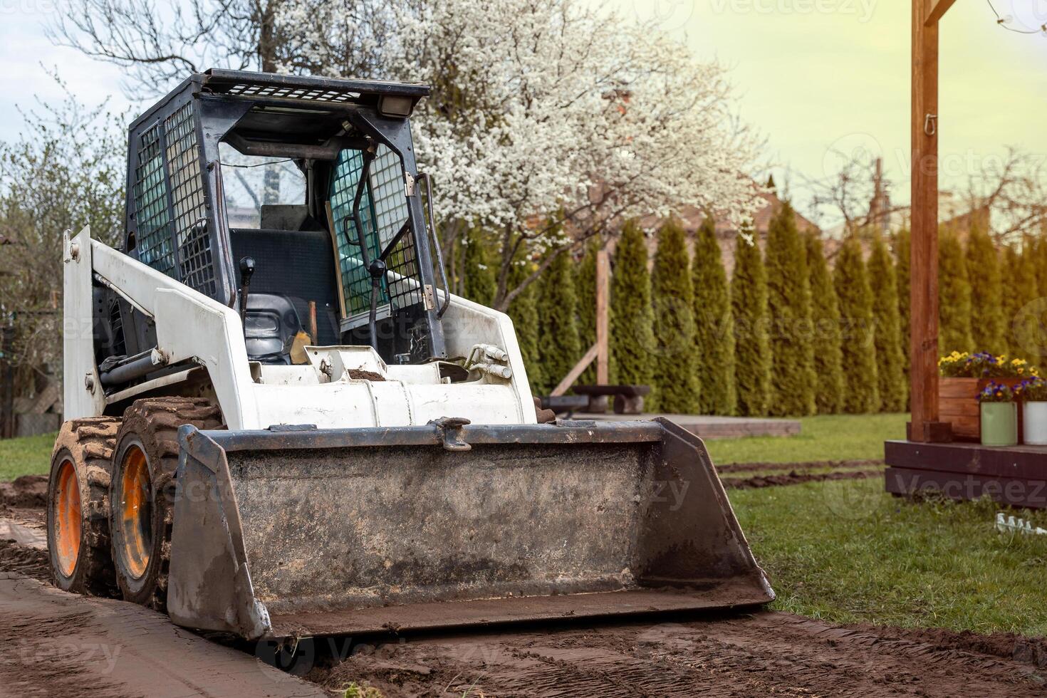 A skid steer loader with a front bucket sits on a dirt path, surrounded by grass, blooming white trees, tall hedges, and a wooden post with flower pots. photo