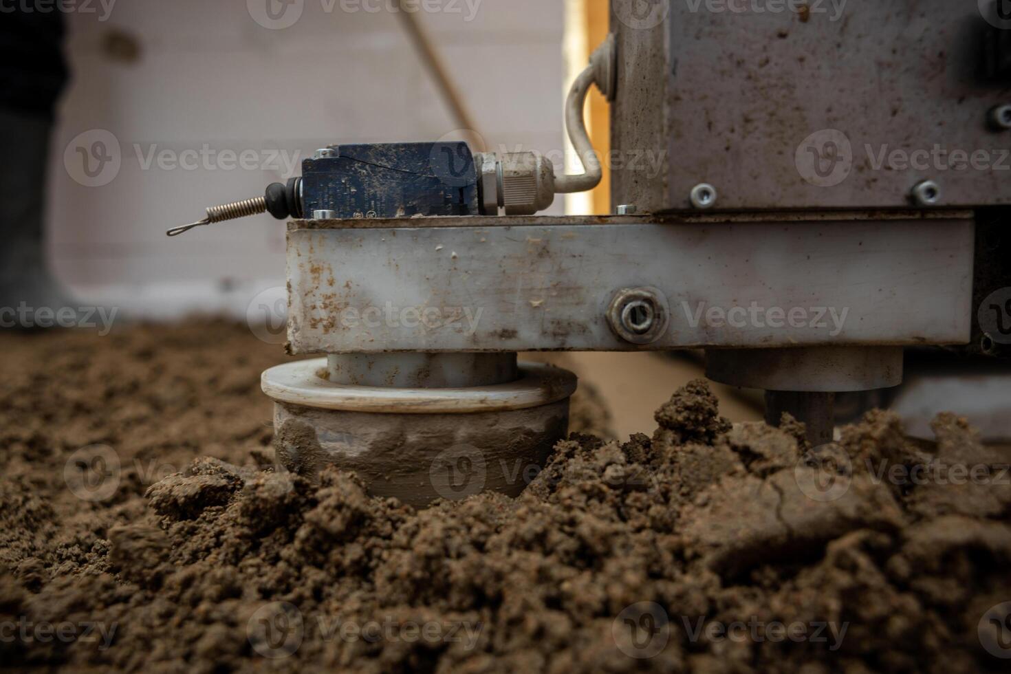 Close up view of construction equipment with a spring loaded mechanism and wiring, set on uneven soil or concrete with a worker's boot visible. photo